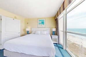 Une chambre d’hôtel avec un grand lit, deux tables de chevet, une fenêtre donnant sur la plage et une porte.