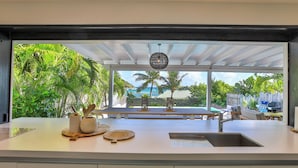 A kitchen with a large island, a sink, and a view of a tropical outdoor area with palm trees and a barbecue grill. - A dream villa for an unforgettable vacation! (Cul-de-Sac)