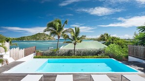 A rectangular swimming pool with a clear blue water surface, surrounded by wooden decking and white lounge chairs. In the background, there is a view of a tropical beach with palm trees and a hilly landscape. - A dream villa for an unforgettable vacation! (Cul-de-Sac)