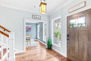 Un couloir avec un plancher en bois, un escalier blanc, une porte avec une fenêtre et une pancarte de bienvenue.