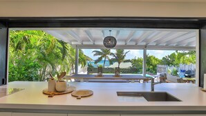 A kitchen with a large island, a sink, and a view of a tropical outdoor area with palm trees and a barbecue grill. - A dream villa for an unforgettable holiday! (SAINT MARTIN)