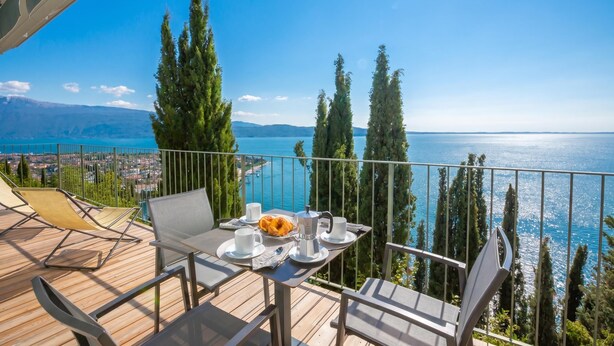Un balcón con una mesa lista para dos personas, con vista a un cuerpo de agua y un paisaje urbano.