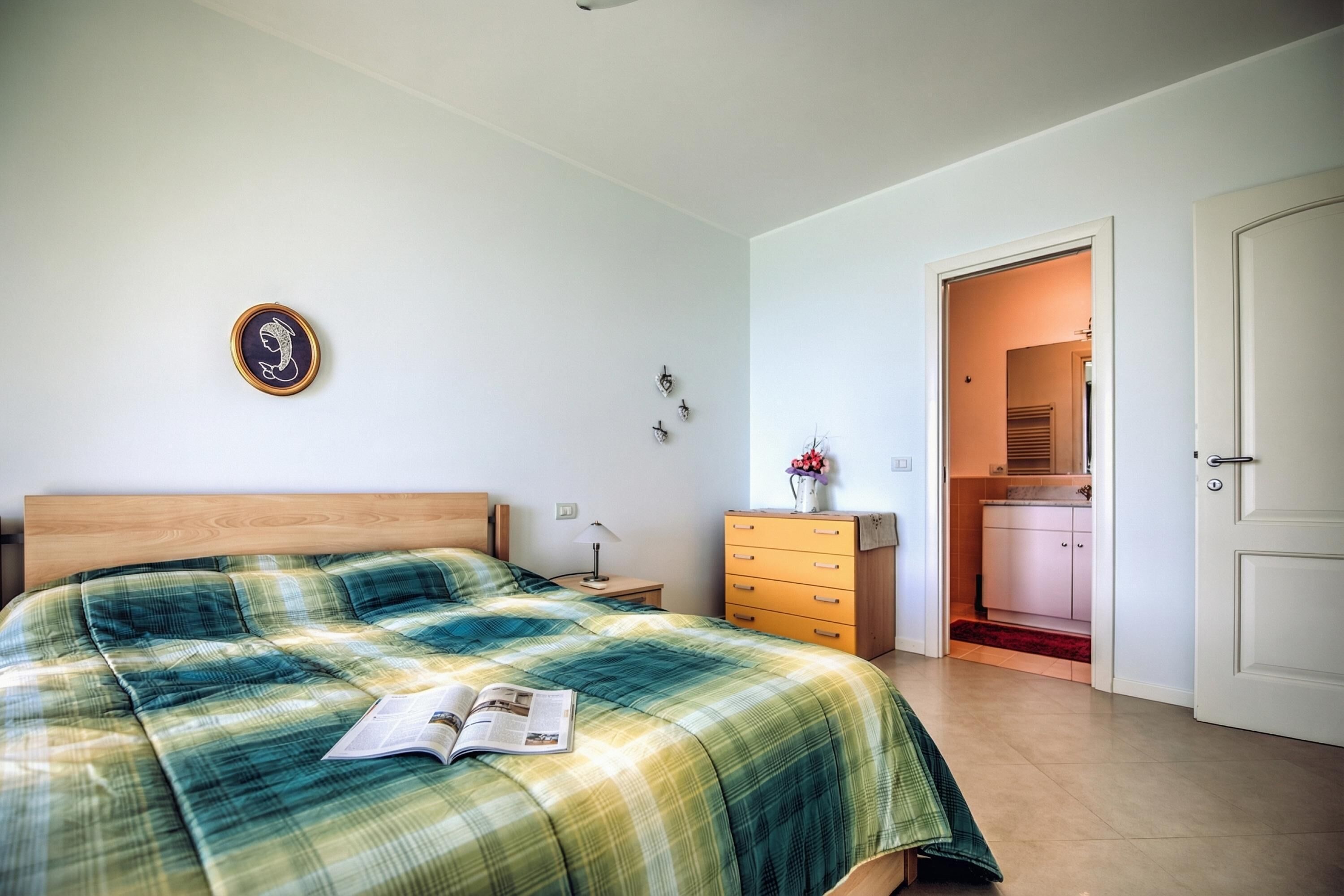 A bedroom with a bed, a dresser, a nightstand, a wall-mounted light, and a door leading to a bathroom.