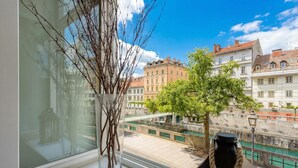 A window with a vase of branches, a lamp, and a view of buildings and a canal. - Tromostovje In Heart Of Ljubljana (Ljubljana)