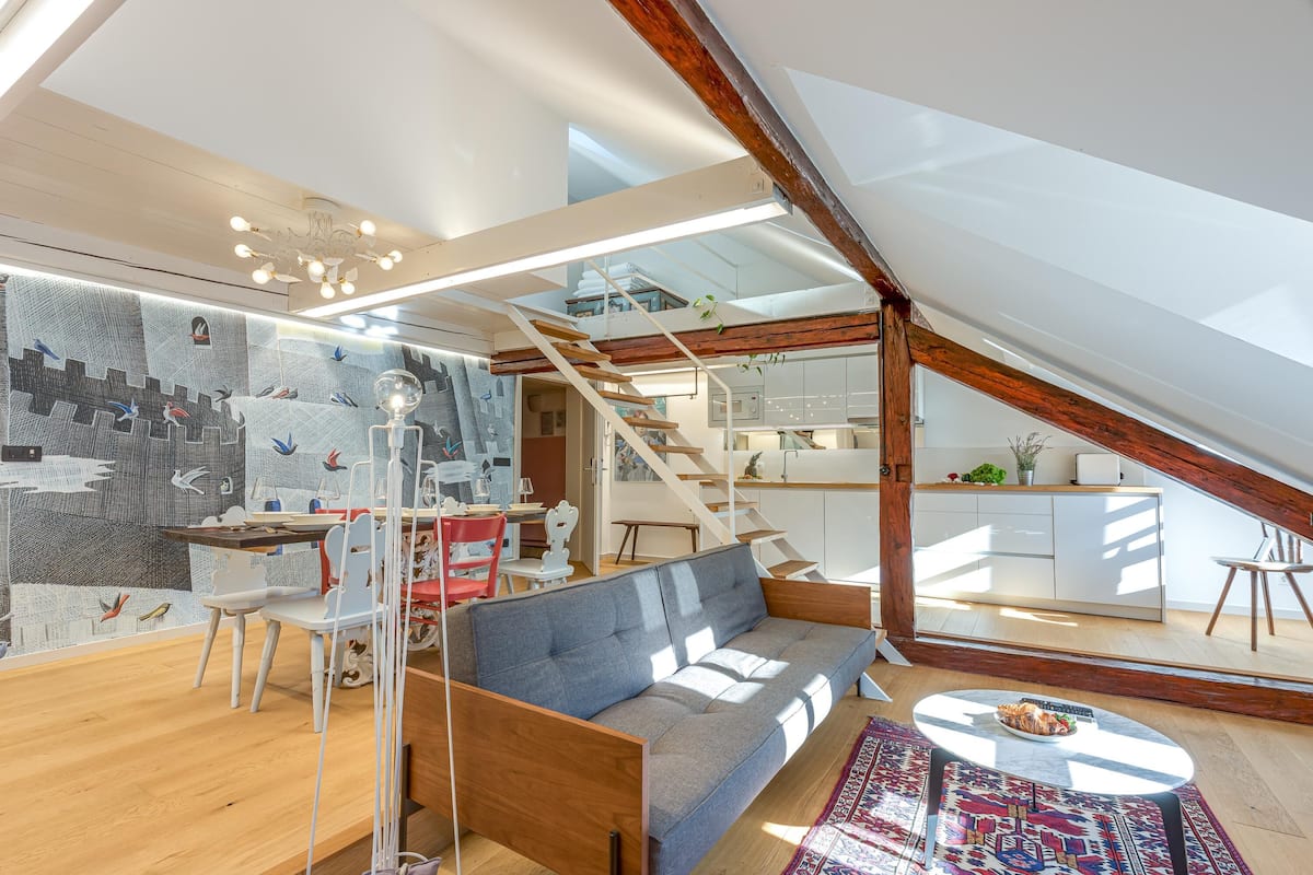 Uma área de estar estilo loft com sofá, mesa de jantar, cadeiras, escada e cozinha ao fundo.