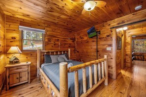 A wooden cabin bedroom with a bed, nightstand, lamp, and a television on the wall.