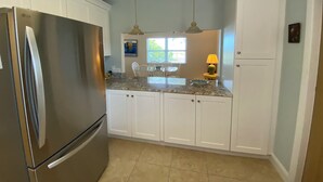A kitchen with white cabinets, a stainless steel refrigerator, granite countertops, pendant lights, and a window with blinds. - Gulf Front Vacation Rental- Anna Maria Island, Florida (Anna Maria)
