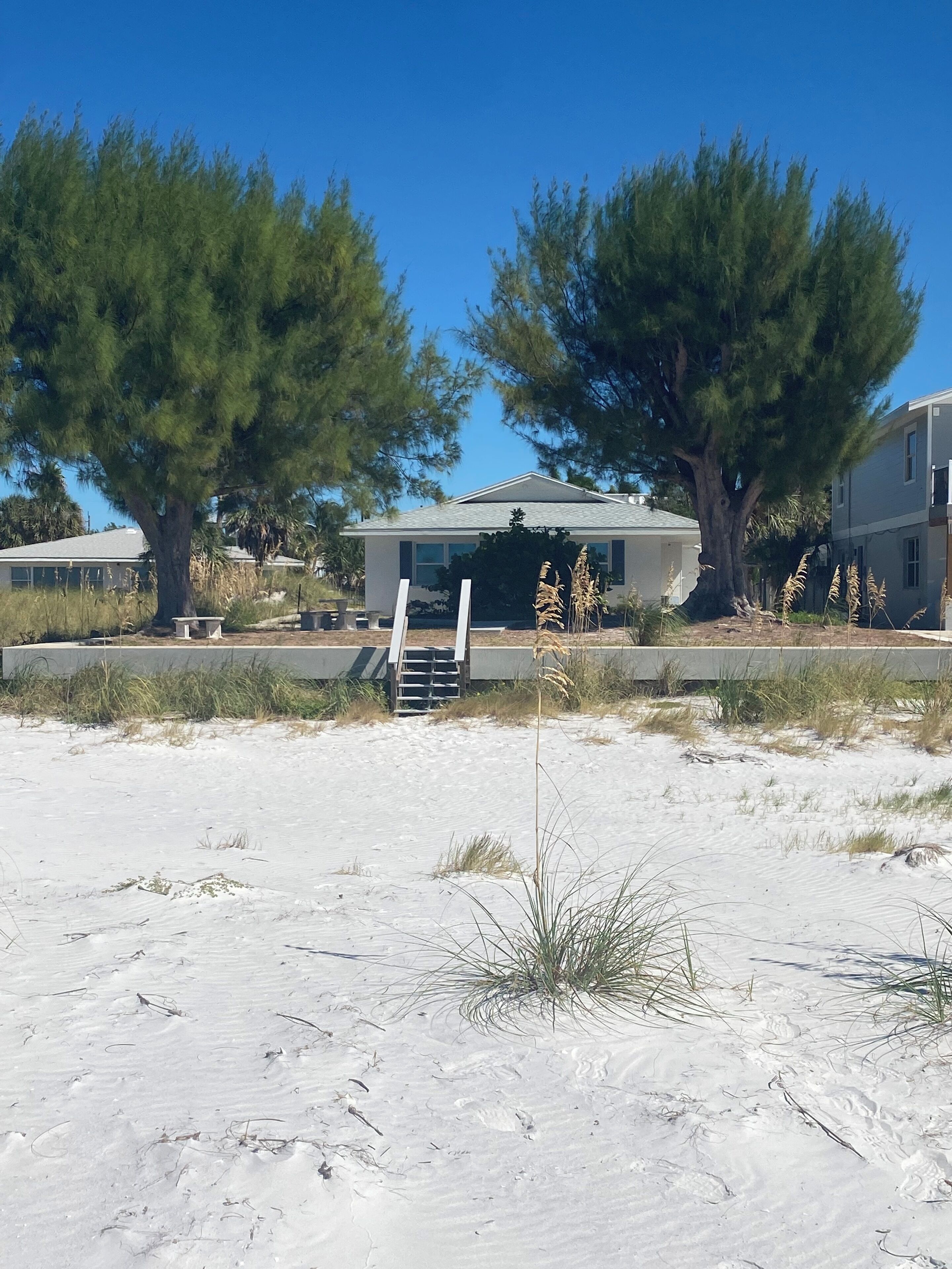 A house with a covered porch, surrounded by sand and sparse vegetation.