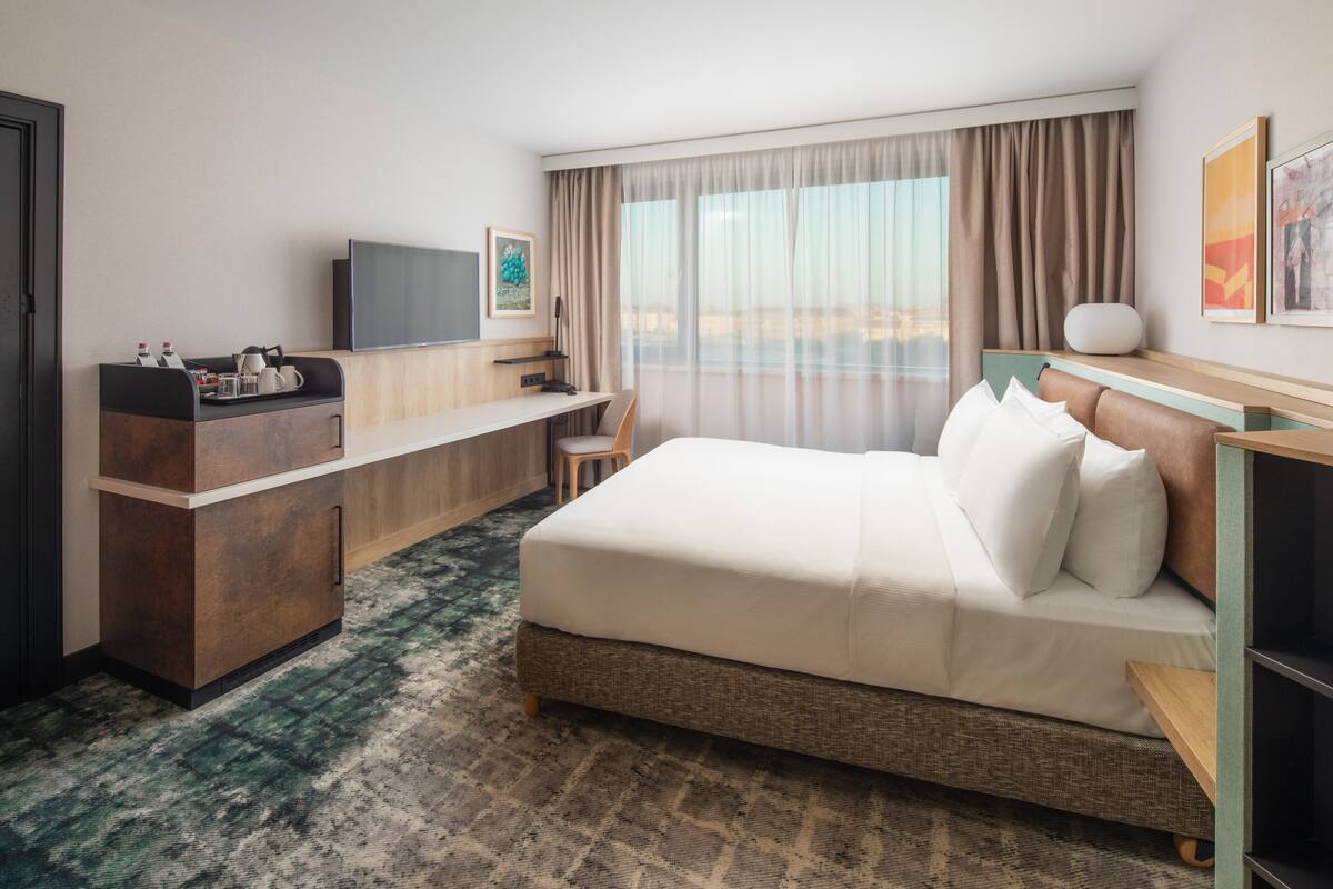 A hotel room with a bed, desk, TV, and a view of the city.