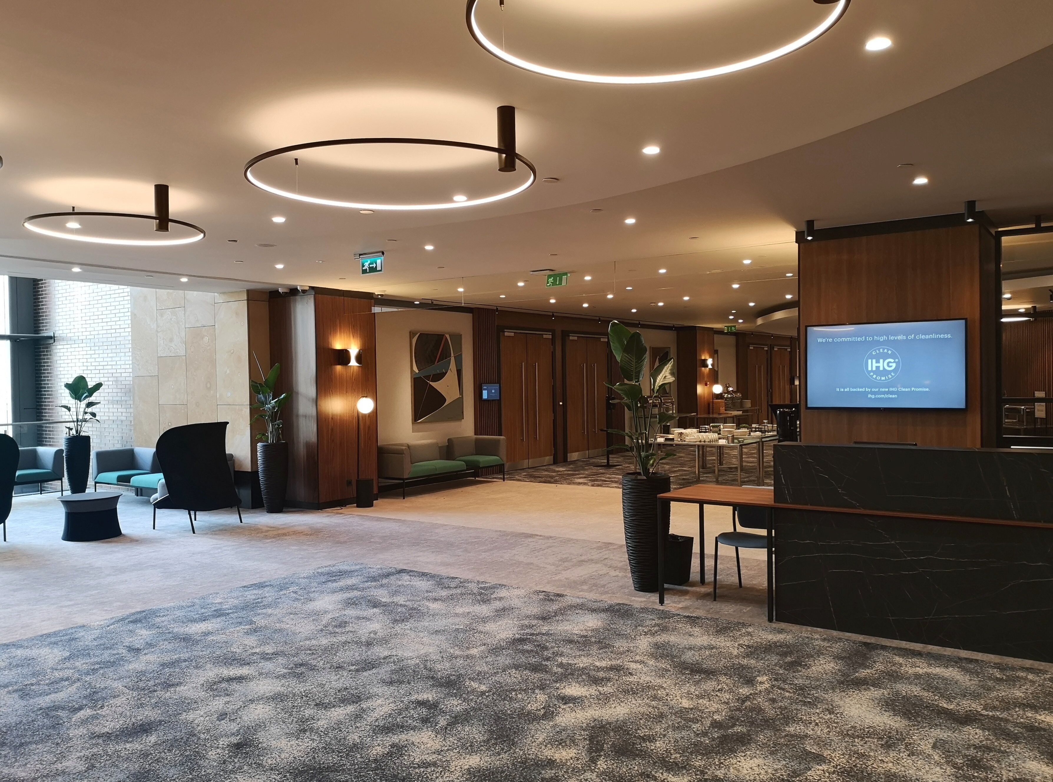 A modern hotel lobby with a reception desk, seating area, and a large screen displaying a cleanliness message.