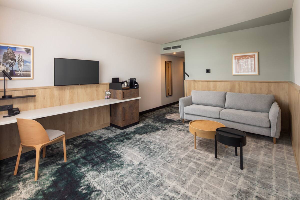 A modern hotel room with a sofa, a desk, a chair, a coffee table, and artwork on the walls.