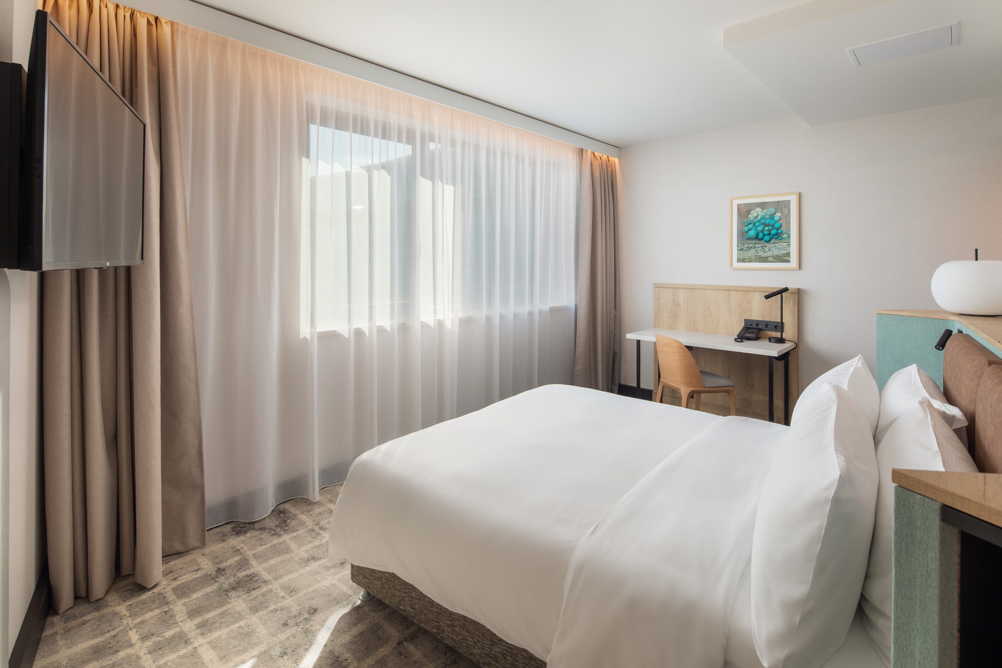 A hotel room with a large bed, a desk, a chair, and a television on the wall.