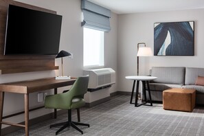 Quarto de hotel com televisão de tela plana, uma mesa com cadeira, sofá, mesa redonda pequena e abajur.