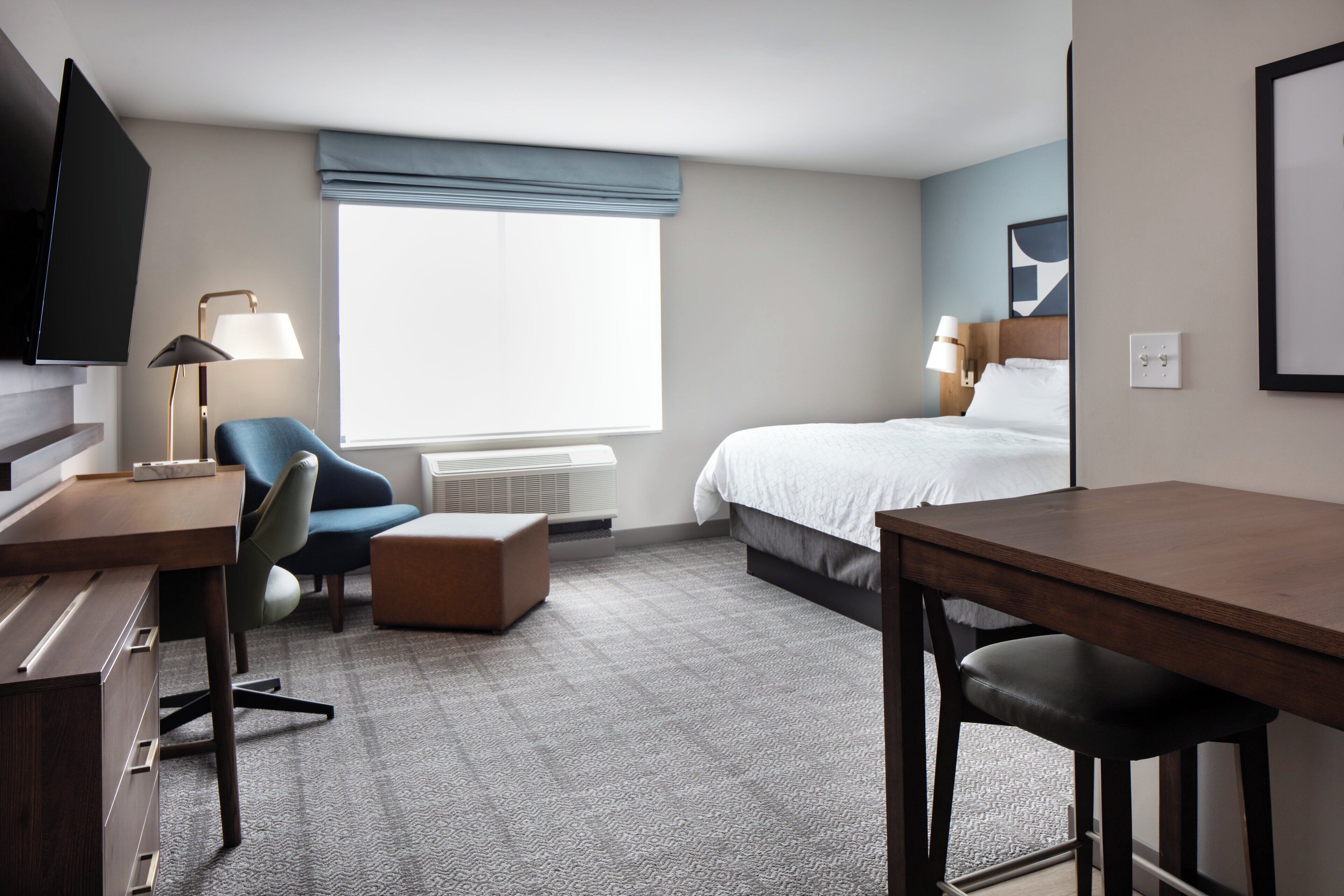 A hotel room with a bed, desk, chair, ottoman, and a television.