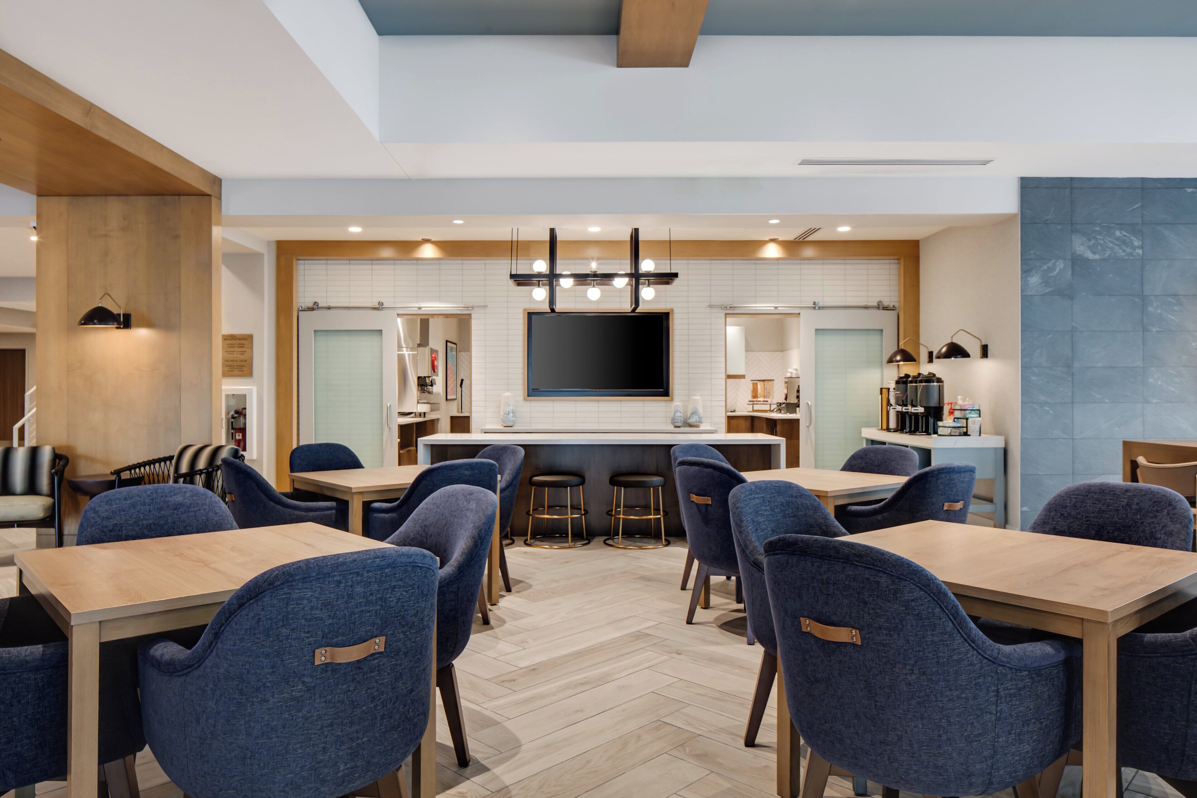 A modern dining area with a central bar, multiple seating arrangements, and a large flat-screen TV mounted on the wall.