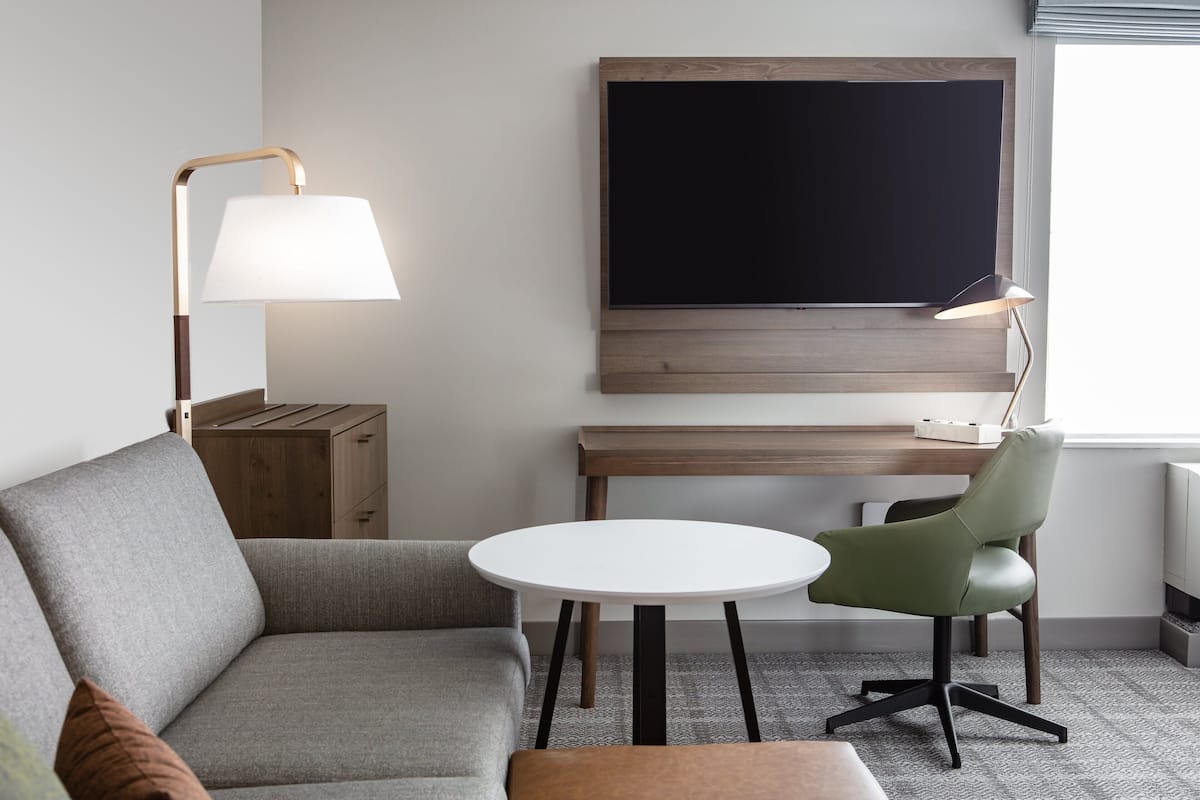 Une chambre d’hôtel moderne avec un canapé gris, une table ronde blanche, une chaise verte, un bureau en bois et un grand téléviseur à écran plat fixé au mur.