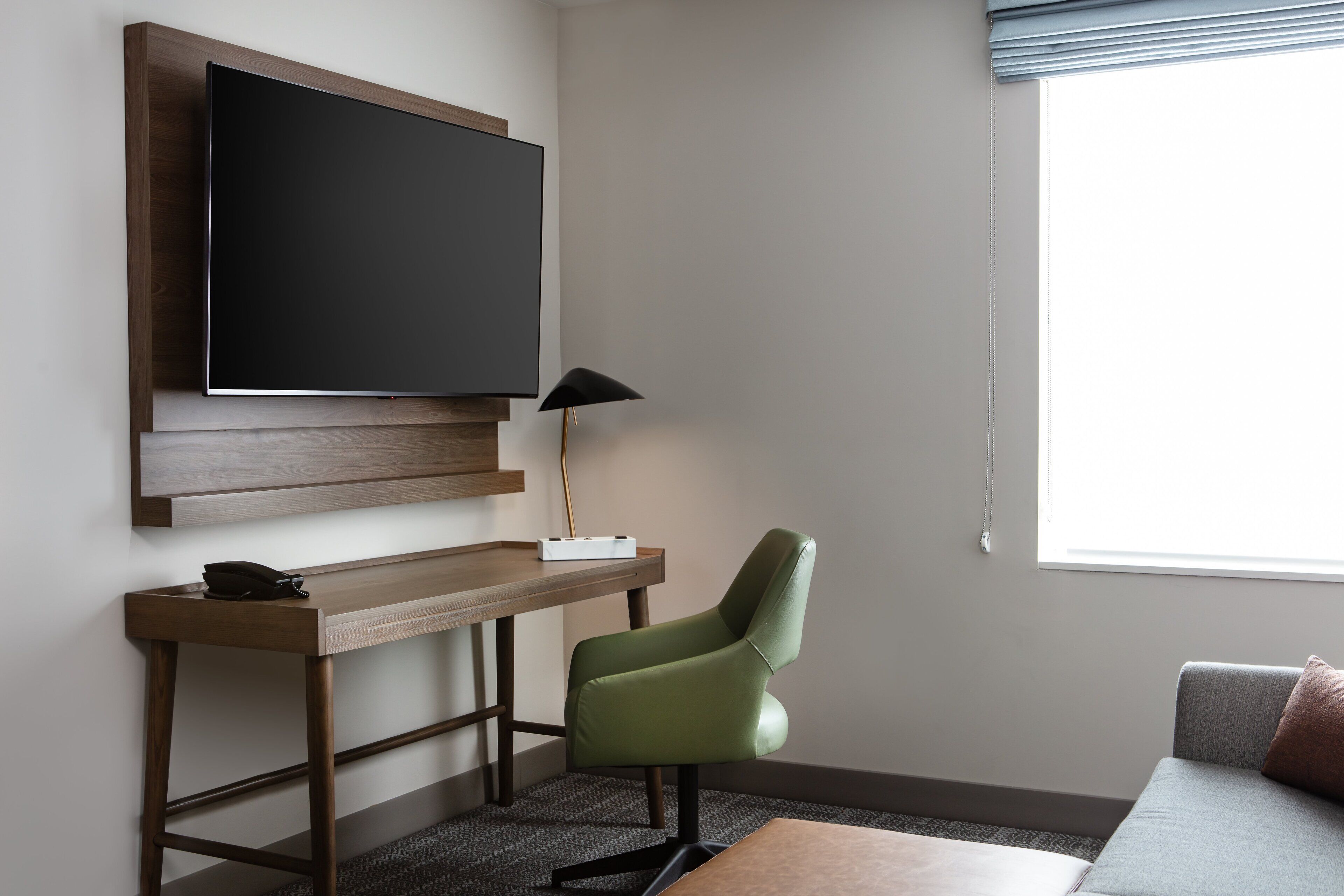 A modern room with a flat-screen TV mounted on a wooden panel, a desk with a lamp, a green chair, and a sofa with a cushion.
