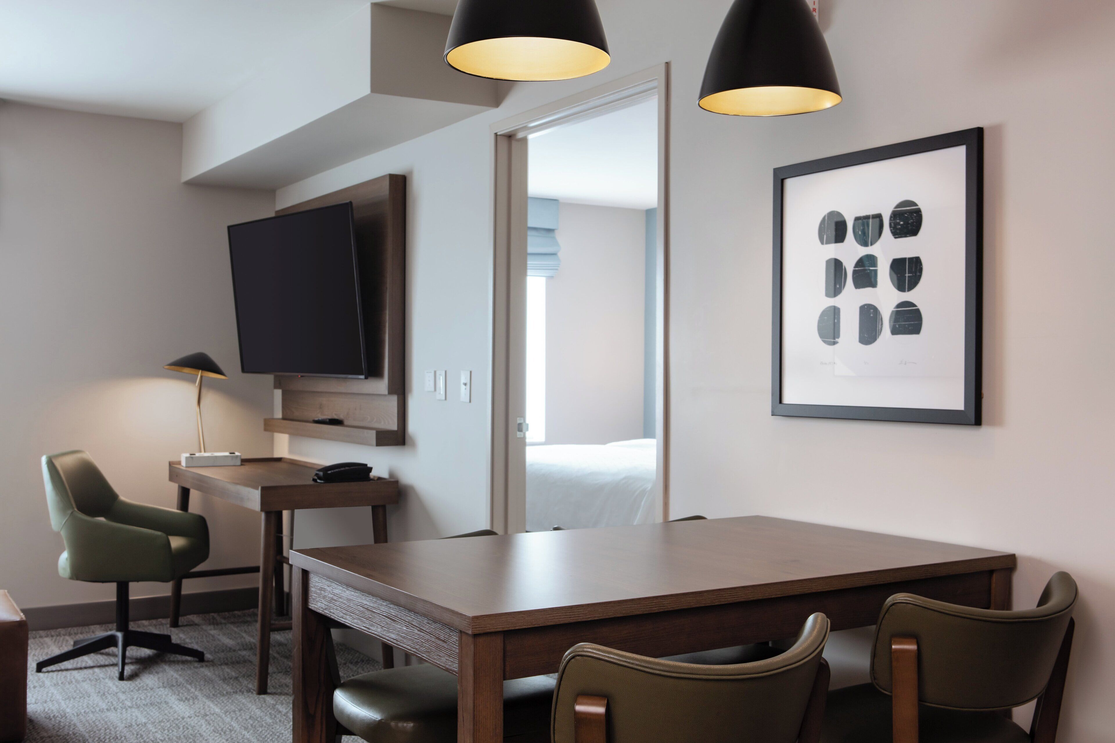 A hotel room with a wooden desk, two chairs, a flat-screen TV, a framed artwork, and a bed visible through an open door.