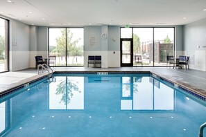 Una piscina con agua cristalina azul, rodeada de sillas y mesas, y grandes ventanales que ofrecen vistas al exterior.
