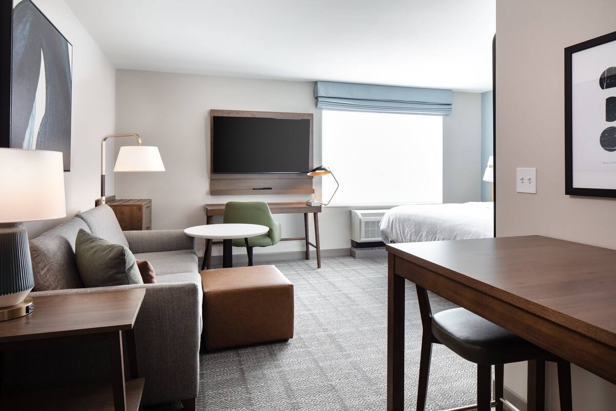 Une chambre d’hôtel équipée d’un canapé, d’une petite table, d’un bureau, d’une chaise, d’un lit, d’une télévision et d’une fenêtre donnant sur un paysage agréable.
