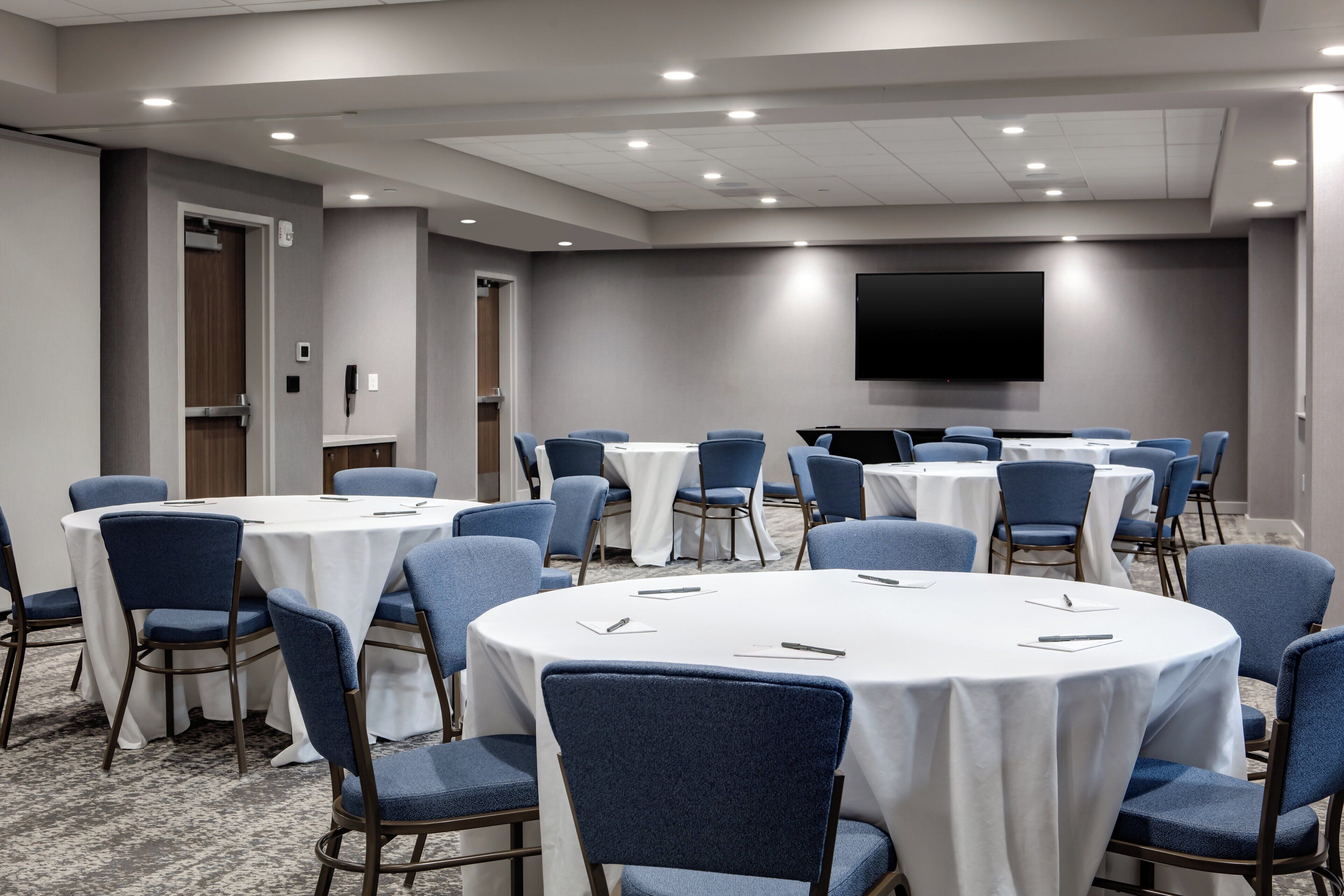 A conference room with multiple round tables, blue chairs, a flat-screen TV mounted on the wall, and a carpeted floor.