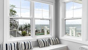 A room with large windows overlooking a coastal area, featuring a white bench with striped cushions. - Expansive Bayfront Home w/ Heated Saltwater Pool & Dock (Ship Bottom)