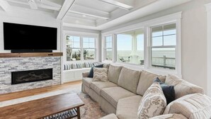 A spacious living room with a large TV, a fireplace, a sofa, and large windows offering a view of the ocean. - Expansive Bayfront Home w/ Heated Saltwater Pool & Dock (Ship Bottom)