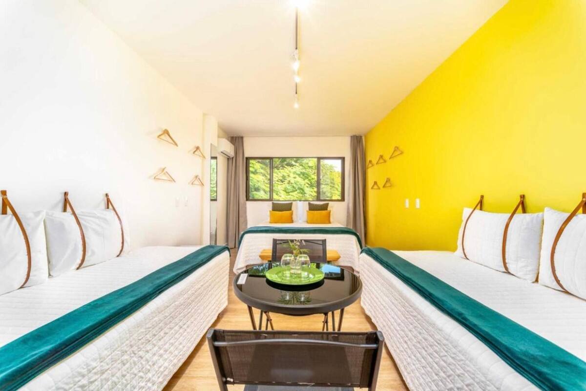 A hotel room with two beds, a dining table, and a view of greenery outside.