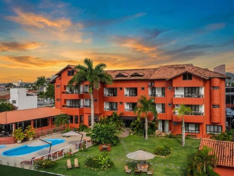 Ein mehrstöckiges Hotel mit Swimmingpool, umgeben von viel Grün und Palmen.