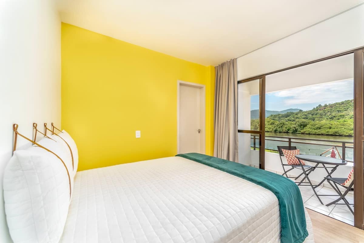 A room with a bed, a balcony with a table and chairs, and a view of greenery.