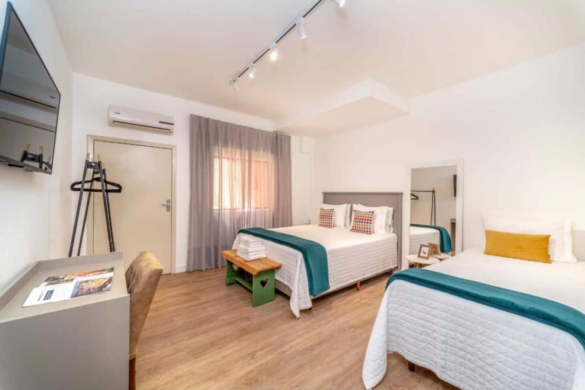 A modern hotel room with a bed, a desk, and a chair. There is a window with curtains, an air conditioning unit, and a mirror on the wall.