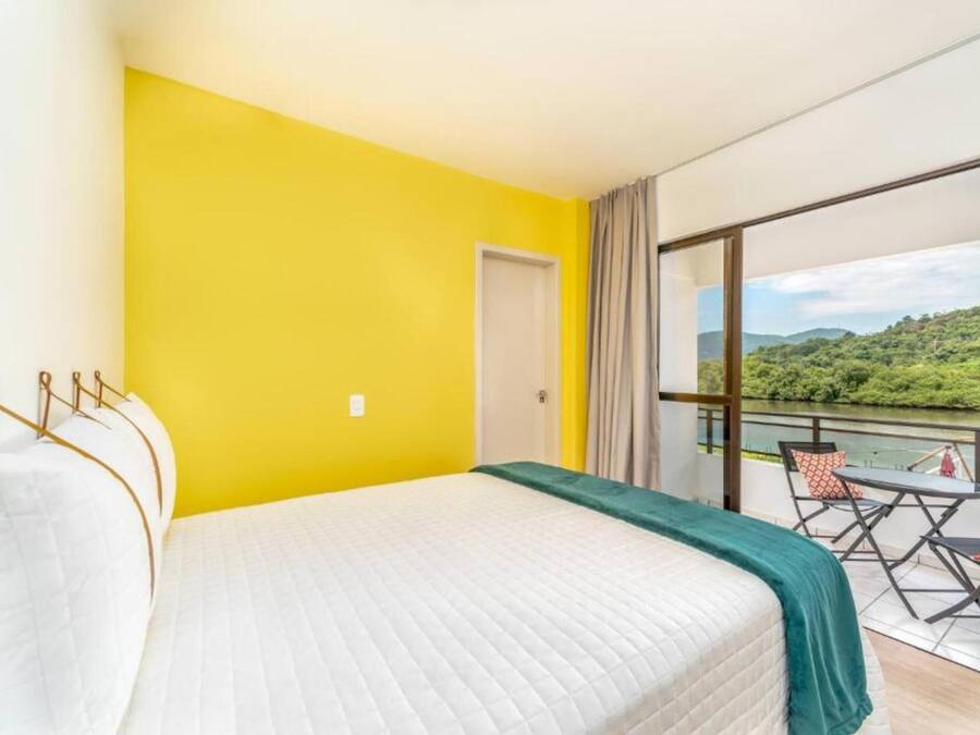 A hotel room with a large bed, a balcony with a table and chairs, and a view of greenery and water.