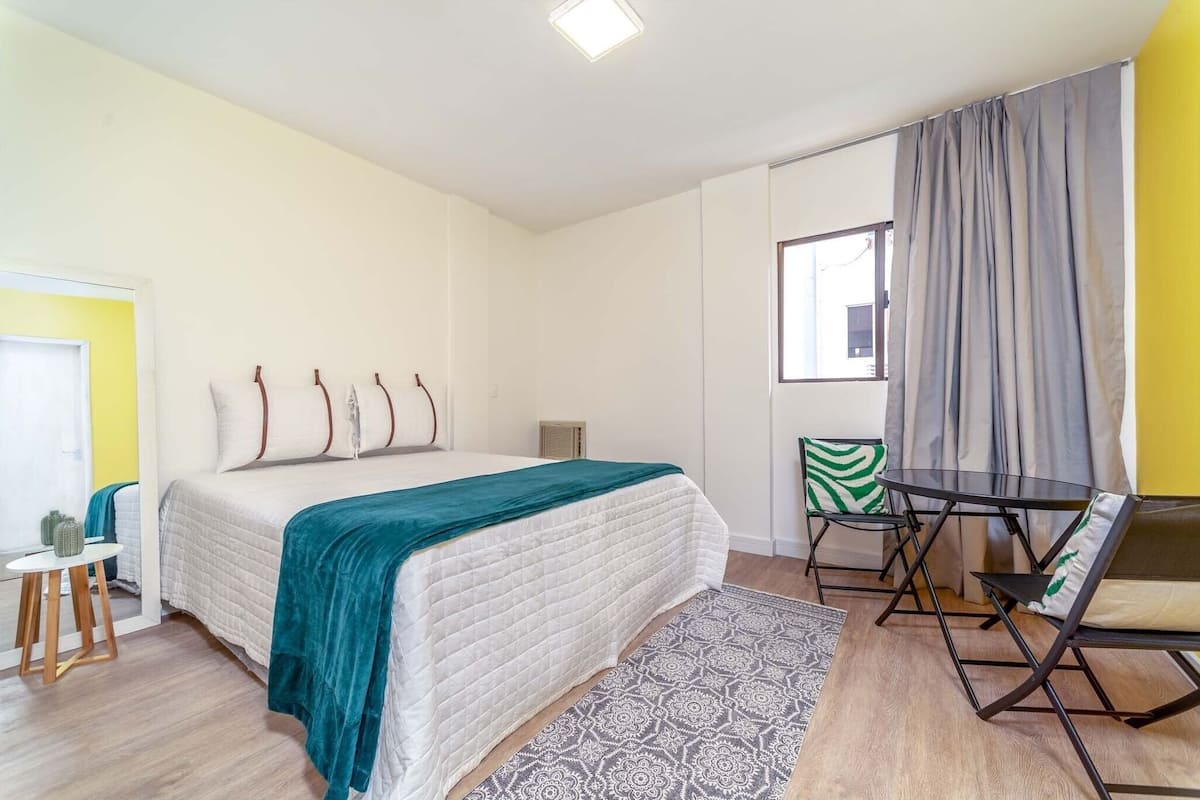 A bedroom with a bed, a small table, a chair, a window, and a curtain.