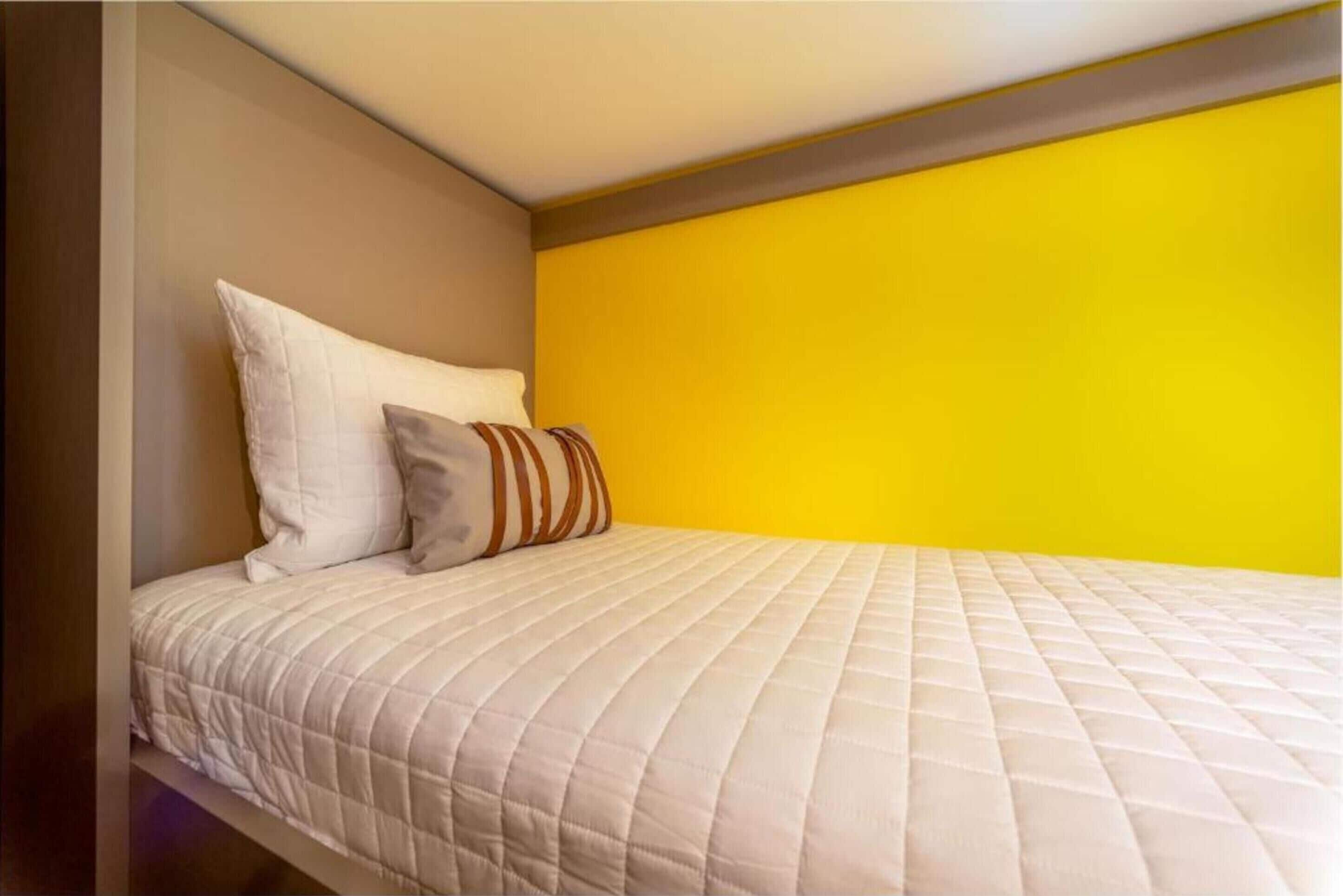 A bed with a quilted white mattress, a pillow, and a striped pillow against a yellow wall.