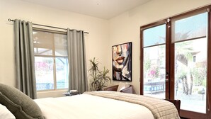 A bedroom with a bed, a window with curtains, a painting on the wall, and a view of the outside.
