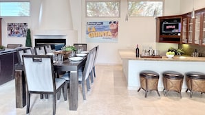 A modern dining area with a long table set for six, surrounded by chairs. There is a kitchen area with a built-in island and bar stools.