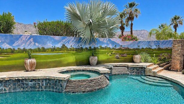 A swimming pool with a stone edge, surrounded by a landscaped garden with palm trees and a mural depicting a golf course.