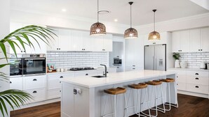 A modern kitchen with white cabinetry, a central island with bar stools, a stainless steel refrigerator, and a built-in oven. - Hamptons Ocean Escape - The most beautiful home for your next luxurious, relaxing getaway (Ulladulla)