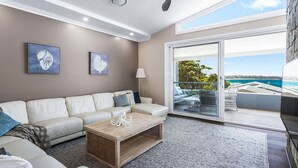 A modern living room with a white sofa, a wooden coffee table, and a large sliding glass door leading to an outdoor balcony with a view of the ocean. - Hamptons Ocean Escape - The most beautiful home for your next luxurious, relaxing getaway (Ulladulla)
