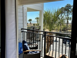 Un balcón con una silla de mimbre y una tabla de surf, con vista a una playa con palmeras y cielo despejado.