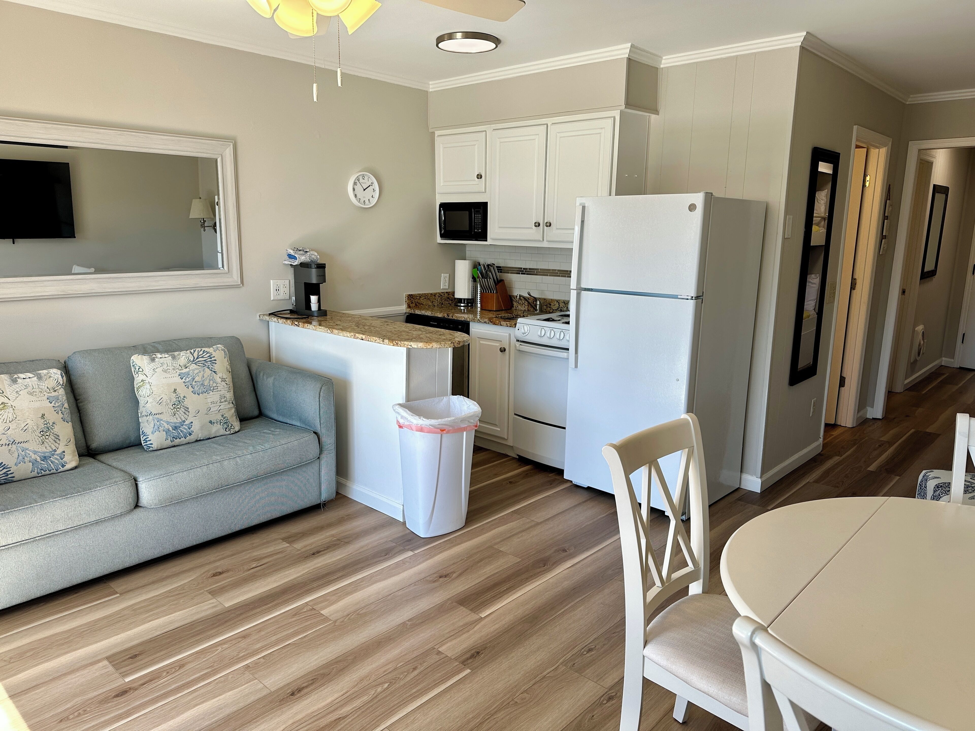A modern kitchen with white cabinets, a refrigerator, a microwave, and a stove. There is a gray sofa with patterned cushions and a small trash bin.