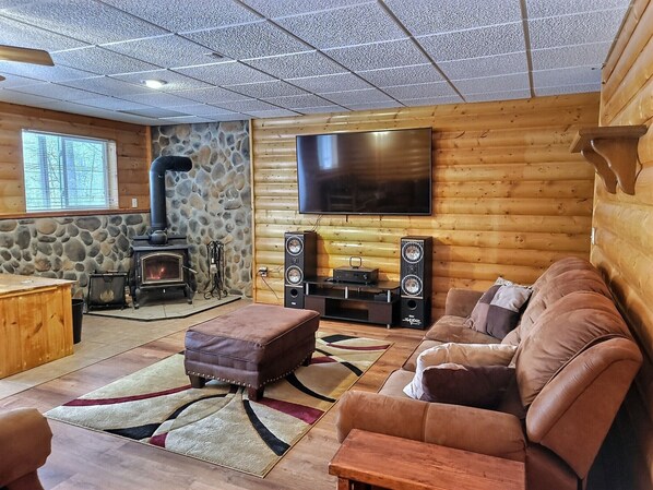 Interior de una cabaña de troncos con chimenea de piedra, un televisor de pantalla plana, un sofá y una mesa de madera.