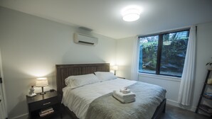A bedroom with a large bed, bedside lamps, a nightstand with books, an air conditioner, and a window with a view of greenery. - Restful, Convenient Getaway Near Downtown and Seattle’s Largest Park (Seattle)