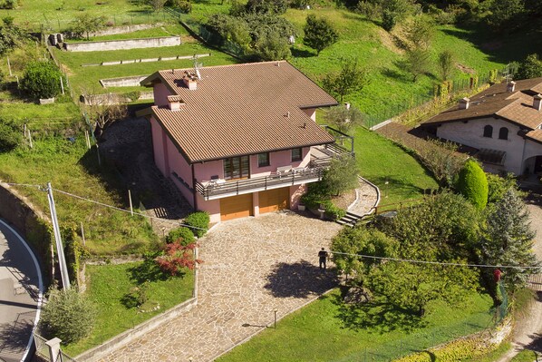A two-story house with a stone-paved courtyard, surrounded by greenery and a fenced area.
