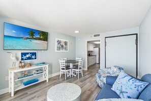 Una sala de estar con comedor, una cocina al fondo y un televisor grande mostrando una escena de playa.