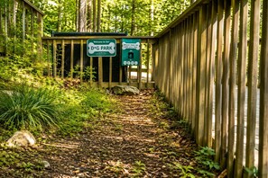 Un sendero de madera conduce a la entrada de un parque, con carteles que indican 'Welcome to Our D*G PARK' y un letrero que dice 'Por favor, limpia después de tu perro'.