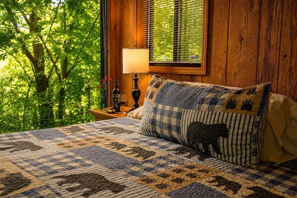 A bedroom with a bed, a nightstand, a lamp, and a window with blinds. - Make Hallmark Memories! Cozy mountain cabin in the woods.  (Gatlinburg)