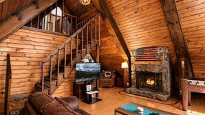 A wooden cabin interior with a stone fireplace, wooden staircase, leather sofa, and a television.