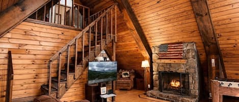 L’intérieur d’un chalet en bois comprend une cheminée en pierre, un escalier en bois, un canapé en cuir et un téléviseur.