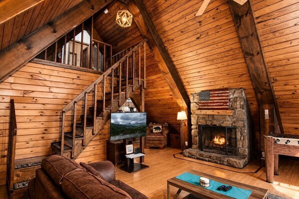 Interior de una cabaña de madera con chimenea de piedra, escalera de madera, sofá de cuero y un televisor.